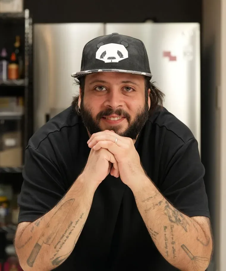 Vini Cordeiro sorrindo, de boné, com as mãos juntas sobre a bancada da cozinha