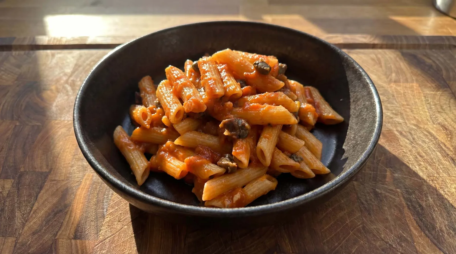 Penne à puttanesca em tigela preta sobre tábua de madeira