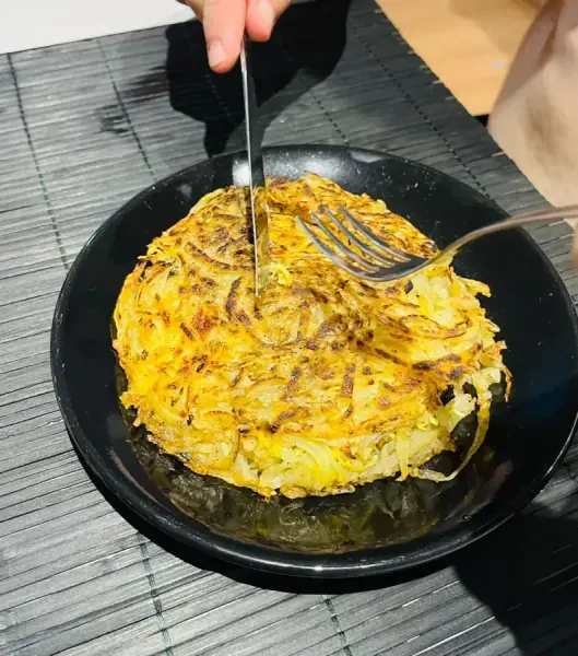 Batata rösti dourada cortada com garfo e faca em prato preto, feita por Rodrigo