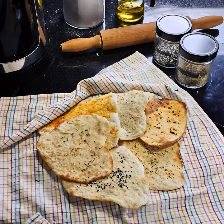 Flatbreads dourados com ervas e gergelim preto sobre pano xadrez, com potes de tempero ao fundo