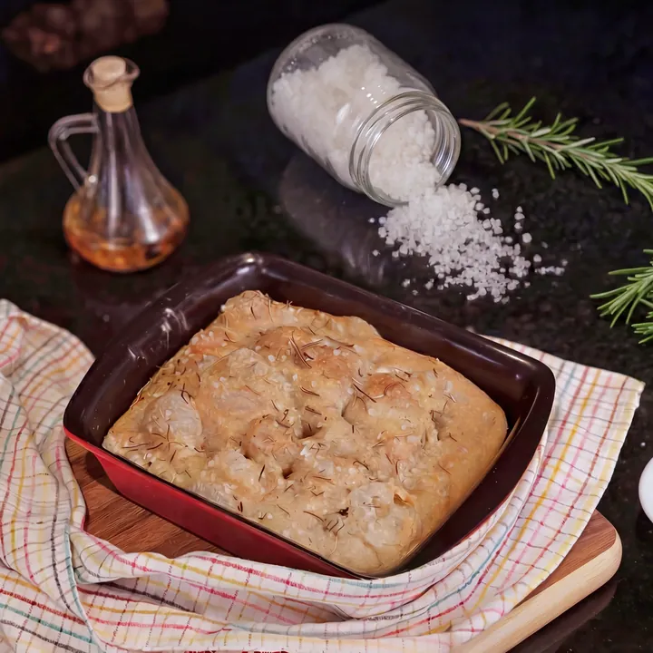Focaccia dourada numa assadeira vermelha sobre pano xadrez, com sal grosso, alecrim e um galheteiro de azeite