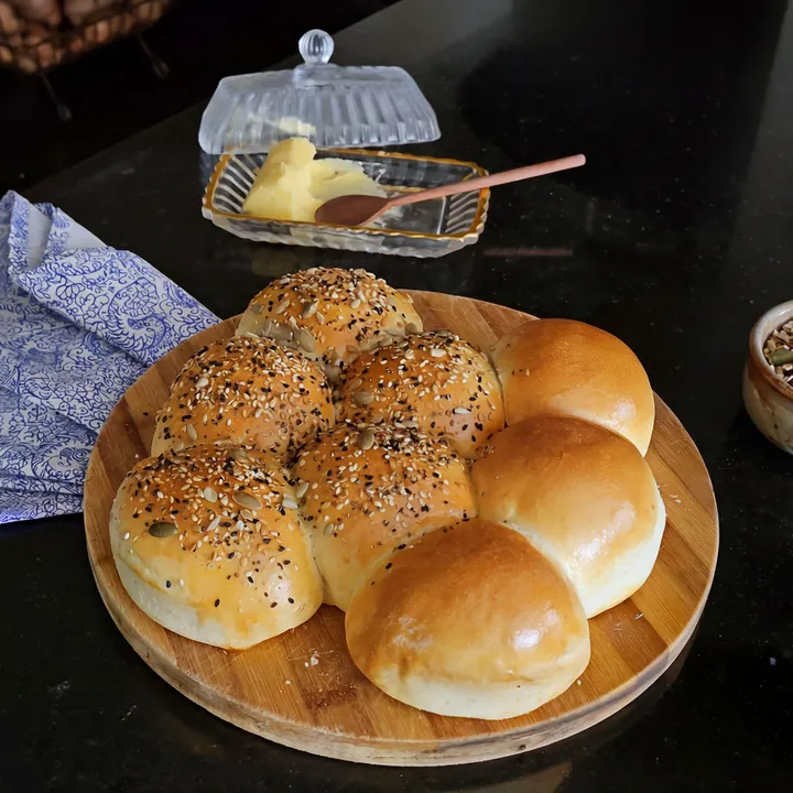 Pão rasga rasga em tábua redonda, metade coberta de sementes, com manteigueira de vidro ao fundo