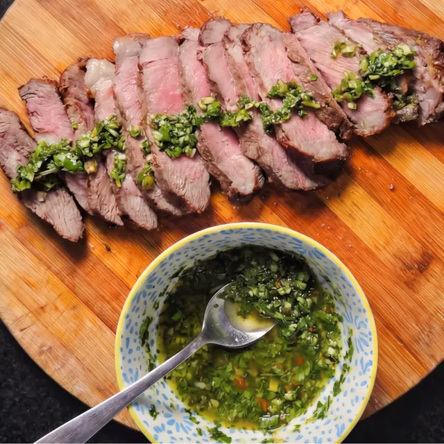 Bife ancho fatiado, rosado por dentro, com chimichurri por cima, em tigela azul sobre tábua de madeira